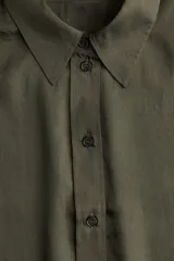 Camisa verde oscuro de manga larga con cuello clásico, cierre frontal con botones y puños abotonados. Confeccionada en tejido satinado con canesú y pliegue en la espalda. Bajo redondeado.