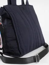 Bolso tipo shopper acolchado de color azul marino, con costuras verticales onduladas en contraste de color azul más claro. Cuenta con asas de mano y una correa ajustable y desmontable de cinta negra con herrajes metálicos plateados.