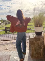 Campera bomber de jean color terracota, con corte holgado. Presenta apliques de cuero cortados y bordados a mano en la espalda, delantera y cuello, formando un diseño de montañas en tonos beige, rosa y verde militar.