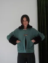 Campera corta color verde azulado con cuello y puños de piel sintética marrón. Cierre frontal con broches tipo toggle de madera.