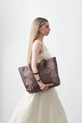Cartera tipo tote bag en símil cuero color marrón oscuro, con bordados decorativos en blanco que evocan un estilo western. Cuenta con asas largas para llevar al hombro.