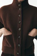 Chaqueta de punto marrón con cuello alto y manga larga. Tiene cierre frontal con botones y bolsillos de parche en el delantero.