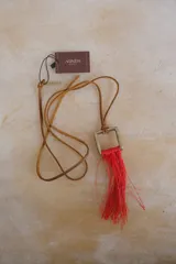 Collar con cordón de cuero marrón y dije cuadrado de metal con flecos rojos.
