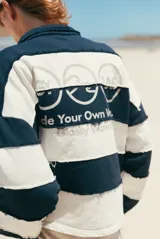 Buzo de tejido de punto grueso, estilo rugby, con cuello polo y tapeta de botones. Diseño a rayas horizontales anchas en blanco y azul marino, con bordes deshilachados en las uniones de las franjas. Presenta un logo bordado en el pecho.