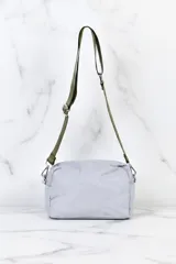 Bolso tipo bandolera o cartera de hombro, color gris claro, con acolchado de costuras onduladas. Posee una correa ajustable de tela color verde oliva.
