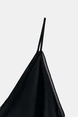 Vestido negro de seda o satén, con escote en V profundo y tirantes finos. Presenta detalles de ribete en terciopelo negro en el escote y un bajo asimétrico con capas superpuestas.