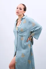 Vestido camisero largo celeste con bordado dorado de hojas, corte holgado, manga larga y cierre frontal con botones.