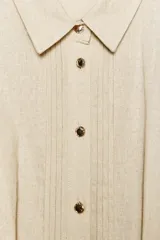 Vestido midi camisero color beige, confeccionado en lino. Presenta cuello solapa, mangas cortas con vuelta, jaretas delanteras, bolsillos laterales ocultos en costura, cierre frontal con botones y cinturón trenzado marrón.
