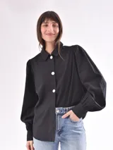 Camisa negra con cuello clásico, abotonada al frente con botones blancos, mangas largas abullonadas y puños con botón. Corte recto.