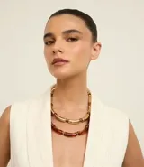 Collar de doble capa con diseño de tubitos curvos. Una de las cadenas presenta tubitos con acabado dorado brillante, mientras que la otra cuenta con tubitos de resina translúcida en tono ámbar, ambos intercalados con pequeñas cuentas metálicas. Posee un cierre de mosquetón y está confeccionado con materiales reciclados.