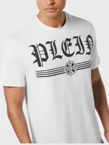Camiseta blanca de manga corta con cuello redondo. Estampado frontal con la palabra "PLEIN" en tipografía gótica negra, flanqueada por dos líneas horizontales y un emblema central con detalles florales y texto pequeño.