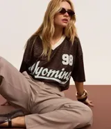 Remera marrón con diseño de damero, cuello en V con borde blanco, mangas cortas y estampado frontal con el número 98 y la palabra "Wyoming" en color celeste.