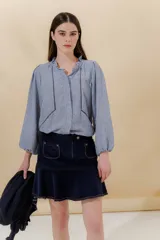 Blusa celeste con cuello mao con volante fruncido y manga larga con puño elástico. En la delantera lleva unas vainillas aplicadas en contratono generando un efecto de dibujo. Se cierra mediante una cartera escondida con botones.