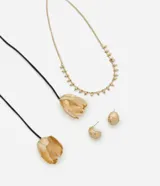 Conjunto de accesorios para mujer compuesto por tres piezas: un collar de cadena fina dorada con pequeñas esferas colgantes, un collar de cordón negro largo con dijes en forma de flor texturizada en los extremos, y un par de caravanas tipo argolla pequeña de diseño grueso. Todas las piezas metálicas presentan un acabado en baño dorado.