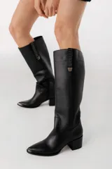 Botas bucaneras de cuero negro con puntera fina y taco cuadrado bajo. Presentan una tira de cuero vertical con apliques metálicos y hebilla decorativa en la parte superior de la caña.