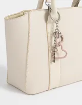 Cartera tipo tote color blanco crudo, con doble asa corta y correa larga regulable y desmontable. Incluye charm colgante con forma de corazón y cierre superior.