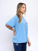 Remera blanca de corte clásico con cuello redondo y mangas cortas con puños tricolor (azul, blanco y rojo). Presenta el logo de Umbro bordado en el pecho izquierdo y el escudo del Club Nacional de Football bordado en el pecho derecho.