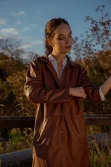 Gabardina marrón de cuero ovino italiano, con cuello camisero, cierre frontal con botones, mangas largas y corte recto.