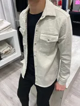 Chaqueta color beige de corte recto, con cuello camisero, cierre frontal con botones metálicos a presión, dos bolsillos con solapa abotonada en el pecho y mangas largas con puños abotonados.