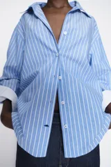 Camisa celeste con rayas verticales blancas, de corte holgado, cuello solapa, escote en pico y mangas largas con puños abotonados.
