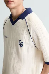 Polo color crema con cuello solapa azul marino, mangas cortas con ribete azul marino, bordado de texto en el pecho y bordado de logo en el abdomen.