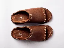 Sandalias de cuero color marrón claro, con punta descubierta y diseño tipo mule. Presentan tachas doradas en el borde superior y suela plana con pequeño tacón. Fabricadas en Uruguay.