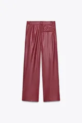 Pantalón de cuero sintético color bordó, de tiro medio, corte recto y cintura cruzada con trabillas. Cuenta con bolsillos laterales, pinzas delanteras y cierre frontal con cremallera, botón y gancho metálico.