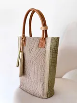Bolso tote de cuero color taupe con textura croco y detalles en verde. Tiene doble asa de mano de cuero marrón y un dije de cuero con flecos. No tiene división interna.