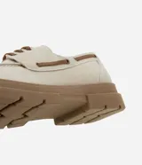 Zapato estilo dockside de color beige claro, confeccionado en gamuza sintética, con cordones marrones y suela track gruesa de goma color marrón. Presenta costuras visibles en la puntera y ojales metálicos para los cordones.