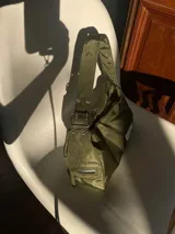 Cartera negra de lona semi impermeable con cierre metálico, forrería de gross y bolsillo interno. Tiene tira regulable y etiqueta blanca con texto en el frente.