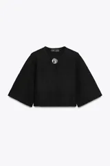 Chaqueta corta negra de tejido texturizado, con cuello redondo y manga ancha tipo kimono que termina por debajo del codo. Cierre frontal con un único botón joya metálico plateado con relieve.