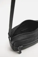 Bandolera negra de acabado engomado con correa ajustable y extraíble. Tiene un compartimento principal con cierre de cremallera, un bolsillo interior con cremallera y un bolsillo frontal con cremallera.