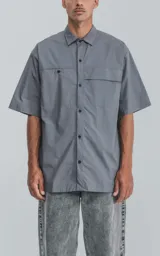 Camisa gris de manga corta confeccionada en poplin 100% algodón, con silueta oversize y dos bolsillos delanteros, uno con solapa y otro con ojal y botón.