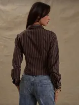 Camisa de manga larga color marrón oscuro con finas rayas verticales blancas. Presenta un corte entallado y ligeramente cropped, con cuello camisero y cierre frontal de botones.