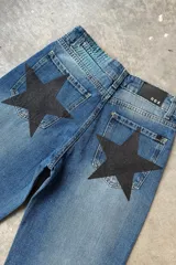 Jean de mezclilla azul medio con efecto desgastado y apliques de estrellas negras de tela texturizada en los bolsillos traseros. Presenta etiqueta de marca negra con texto blanco en la cintura trasera.