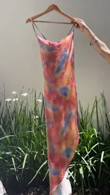 Vestido midi con estampado tie-dye en tonos rosados, azules y beige. Tiene escote drapeado, breteles finos y falda irregular con abertura lateral.