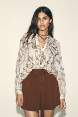 Camisa semitransparente con estampado animal print en tonos beige y marrón, de cuello redondo y escote pico, con mangas largas abullonadas. Detalle de volantes e hilo metalizado. Cierre frontal con botones.