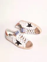 Zapatillas urbanas Golden Goose modelo Super-Star, de cuero blanco con detalles en marrón y estrella negra con glitter en el lateral. Diseño con efecto desgastado en la suela y cordones.
