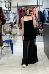 Vestido negro de largo midi, con tirantes finos y escote recto. La falda es asimétrica, con una capa superior de gasa transparente que cae hasta el tobillo y una base más corta de crepé elastizado. Incluye una bufanda larga de gasa a juego que se ata al cuello.