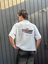 Remera amarilla de corte holgado con estampado trasero de un yate y texto con nombres de ciudades.