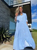 Vestido camisero largo celeste con rayas verticales blancas, cuello camisero, manga larga con puño y abotonadura frontal. Diseño escalonado con volantes en cada corte.
