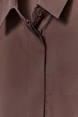 Camisa marrón de popelín con cuello solapa, manga corta con vuelta, bolsillo de plastrón en el pecho, bajo con aberturas laterales, pinzas en la espalda y cierre frontal con botones ocultos.