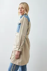 Trench largo color beige con secciones en denim azul, cuello camisero, cierre frontal con botones y bolsillos con solapa en el pecho.