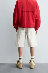 Bermuda de denim color blanco roto con efecto encerado, corte amplio (baggy fit) y cinco bolsillos. Presenta cierre frontal con cremallera y botón.