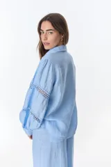 Camisa celeste de lino con cuello clásico, abotonada al frente y mangas largas con detalle de puntilla en los hombros.