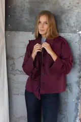 Chaqueta color bordó con cuello camisero, manga 3/4 y cierre frontal con botones negros. Presenta detalle de volantes en el frente y bolsillos de parche.