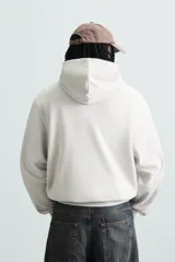 Sudadera con capucha color crudo, de corte relajado, confeccionada en tejido de algodón con interior rizado. Cuenta con cuello con capucha, manga larga, bolsillo frontal tipo canguro y acabados en rib.
