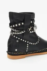 Bota de caña media color negro confeccionada en cuero, con suela de goma y terminaciones de costura manual y remaches metálicos. Presenta un cinto ornamental con tachas, cadenas y piezas en metal.
