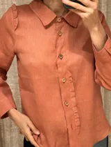 Camisa color ladrillo de lino, corte femenino, con cuello clásico, cierre frontal con botones de madera, mangas largas con puños abotonados y volados en los hombros y en el frente.