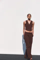 Pantalón marrón de tiro medio con trabillas, bolsillos laterales y de vivo en espalda. Detalle de pliegues y cierre frontal con cremallera, botón interior y gancho metálico.